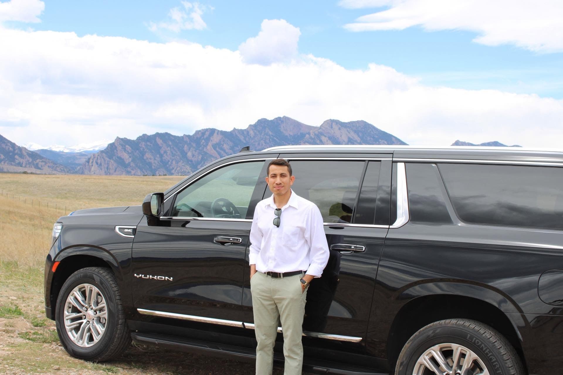 A private driver standing beside a black GMC Yukon with Colorado mountains behind him.